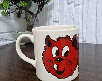 May include: Cream-colored ceramic mug featuring a cartoon red cat face with black accents. The cat has a wide smile and a single tooth. The mug has a curved handle and is suitable for hot beverages.