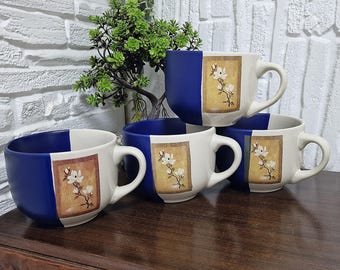 May include: A set of four ceramic mugs. Each mug has a white body with a blue accent and a handle. A rectangular image of a floral design is on the white portion of the mug. The mugs are stacked on a wooden surface.