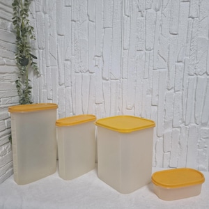 Può includere: Un set di quattro contenitori Tupperware vintage. I contenitori sono traslucidi con coperchi gialli. Le forme variano da rettangolari a quadrate, offrendo versatili opzioni di stoccaggio. I contenitori sono disposti su una superficie bianca.
