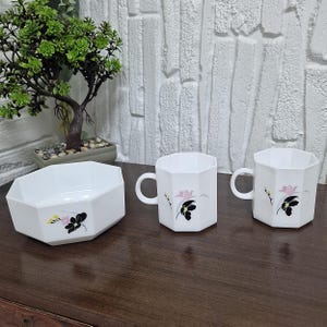 May include: A white, geometric-shaped bowl and two mugs with handles. Each piece features a floral design with pink and black flowers. The set is displayed on a dark wooden surface.