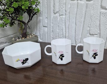 May include: A white, geometric-shaped bowl and two mugs with handles. Each piece features a floral design with pink and black flowers. The set is displayed on a dark wooden surface.