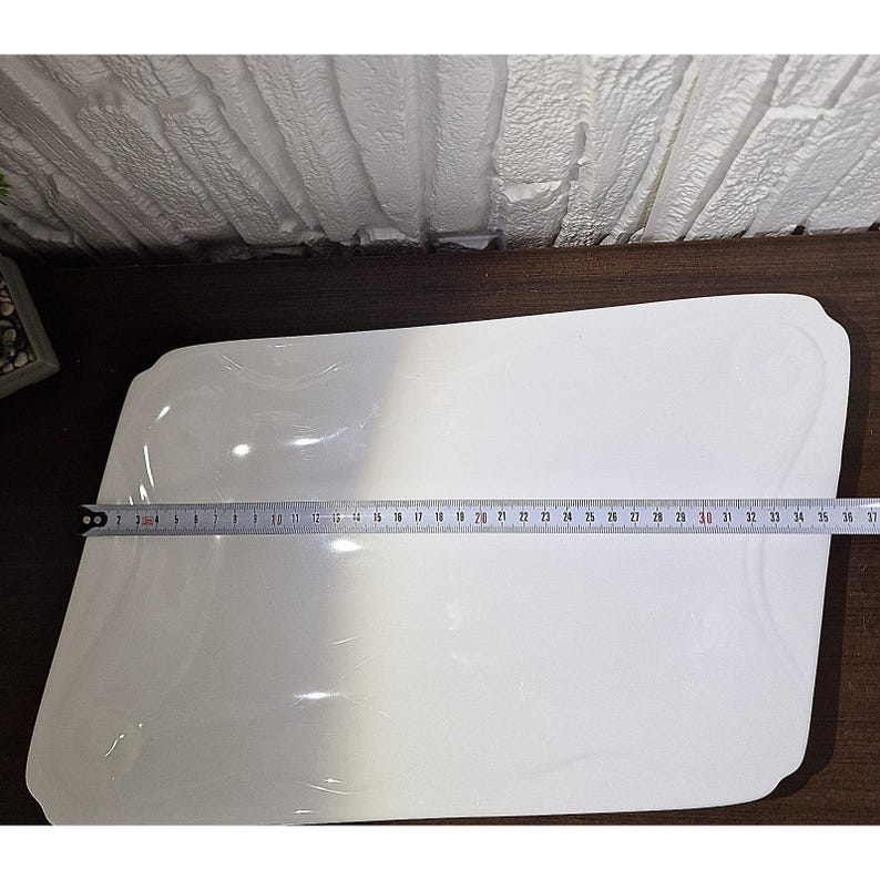 Puede incluir: Una fuente rectangular blanca con bordes suavemente curvados. Una cinta m&eacute;trica muestra que la fuente mide aproximadamente 91 cm de largo. La fuente est&aacute; sobre una superficie de madera oscura, con una pared de ladrillo blanco al fondo.
