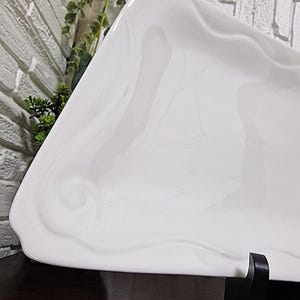 Puede incluir: Una fuente de cer&aacute;mica blanca de forma org&aacute;nica. La superficie brillante tiene patrones sutiles y arremolinados. La fuente se exhibe sobre una superficie oscura, con una vista de una pared de ladrillo y vegetaci&oacute;n en el fondo.