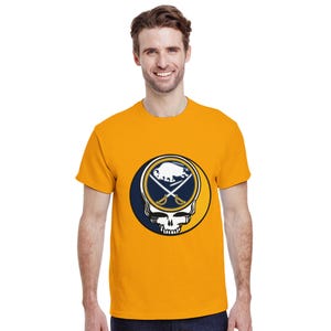 May include: Mustard yellow t-shirt featuring a graphic of a skull with crossed swords and a buffalo head. The design is set within a navy blue and gold circle. The shirt is short-sleeved.