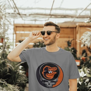 May include: A gray t-shirt with a graphic of a cartoon baseball player's head inside a skull with a black, orange, and white color scheme.