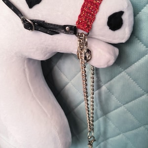 May include: A white plush horse head with black felt eyes and a red beaded halter. A black leather strap and silver chain with a decorative charm are attached to the halter. The horse has a white mane.