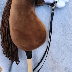 May include: A brown hobby horse with a plush head and a wooden stick. The horse has a black bridle and reins, and a long brown mane. The horse is resting on a light gray surface.