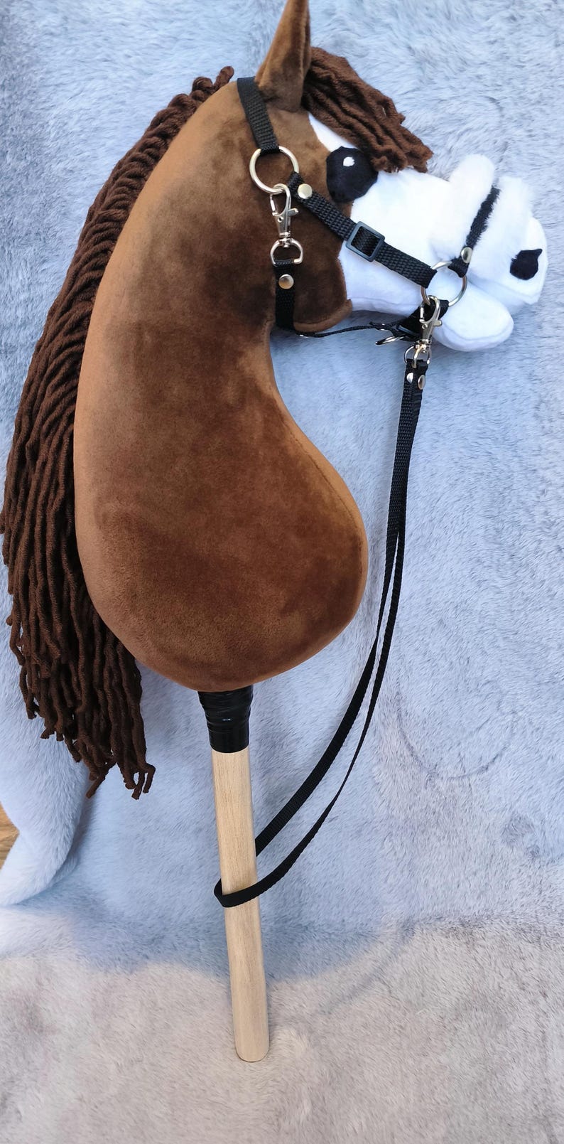 May include: A brown hobby horse with a plush head and a wooden stick. The horse has a black bridle and reins, and a long brown mane. The horse is resting on a light gray surface.