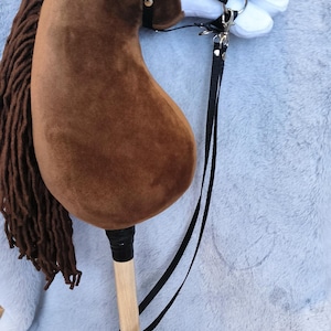 May include: A plush brown hobby horse with a white face and black bridle. The horse has a long brown mane and a wooden stick handle. The toy is designed for children's play and is set against a soft, gray background.