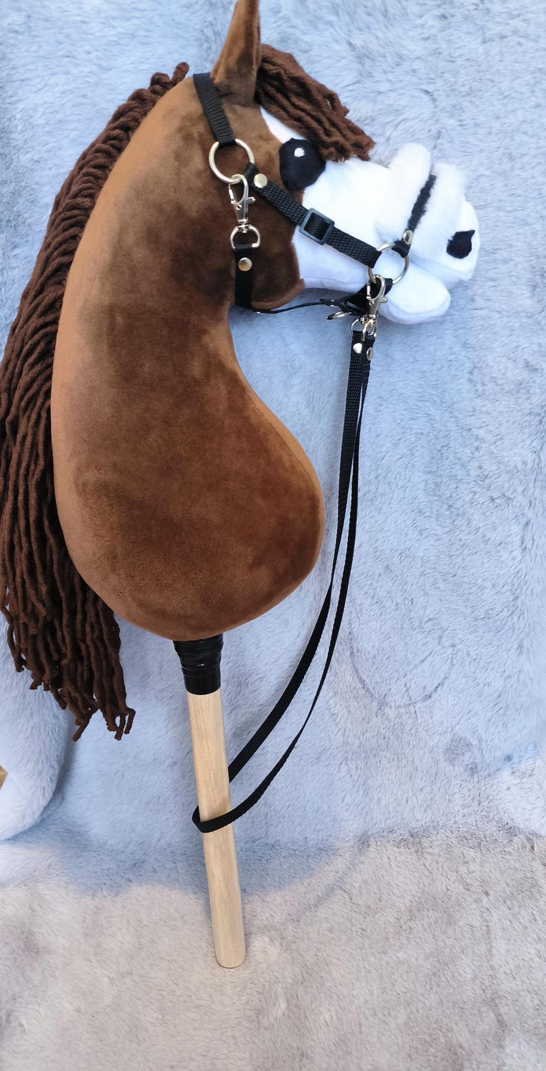 May include: A plush brown hobby horse with a white face and black bridle. The horse has a long brown mane and a wooden stick handle. The toy is designed for children's play and is set against a soft, gray background.