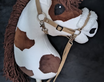 May include: A plush hobby horse with a white body and brown spots, mane, and forelock. It features a tan bridle with metal hardware and a wooden stick handle. The toy is designed to resemble a horse's head.