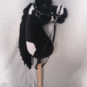 Op de afbeelding: Een zwart-wit stokpaard met een houten stok. De paardenkop heeft een zwarte manen en een zwart halster met goudkleurige ringen. Witte vlekken zitten op het gezicht en het lichaam.