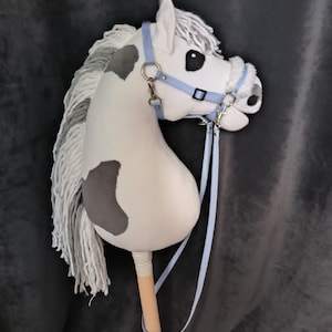 Puede incluir: Un caballo de balancín blanco y gris con una brida y riendas azules. El caballo tiene una larga melena y cola blancas. El caballo está en un palo de madera.