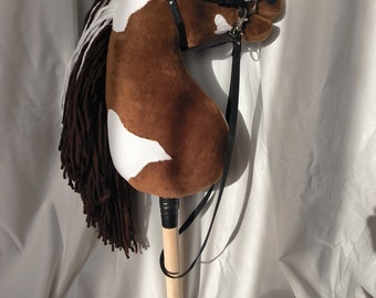 May include: A brown and white pinto horse stick horse toy with a black bridle and reins. The horse has a brown mane and tail made of yarn.