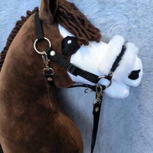 May include: A plush brown hobby horse head with a black bridle and reins. The horse has a white muzzle, black eyes, and a brown mane. The bridle features silver-colored metal hardware.