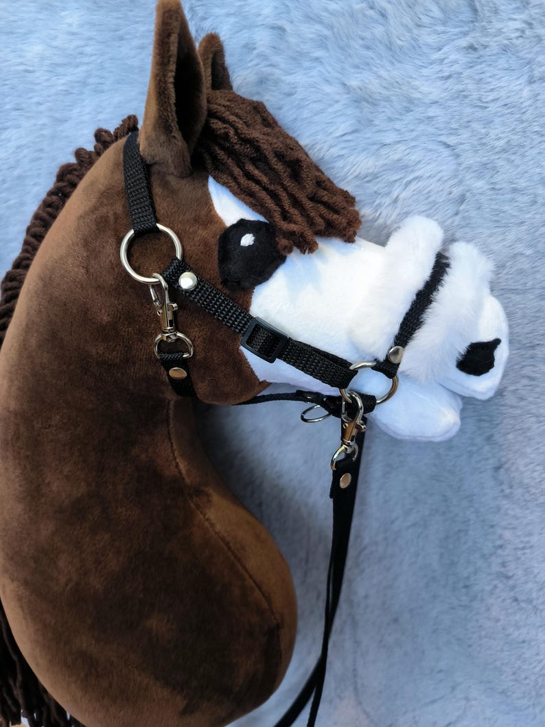 May include: A plush brown hobby horse head with a black bridle and reins. The horse has a white muzzle, black eyes, and a brown mane. The bridle features silver-colored metal hardware.