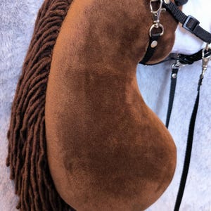May include: A brown hobby horse with a plush head and a long, braided mane. The horse has a black bridle with silver-colored metal hardware and a wooden stick handle. The background is a light gray, textured fabric.