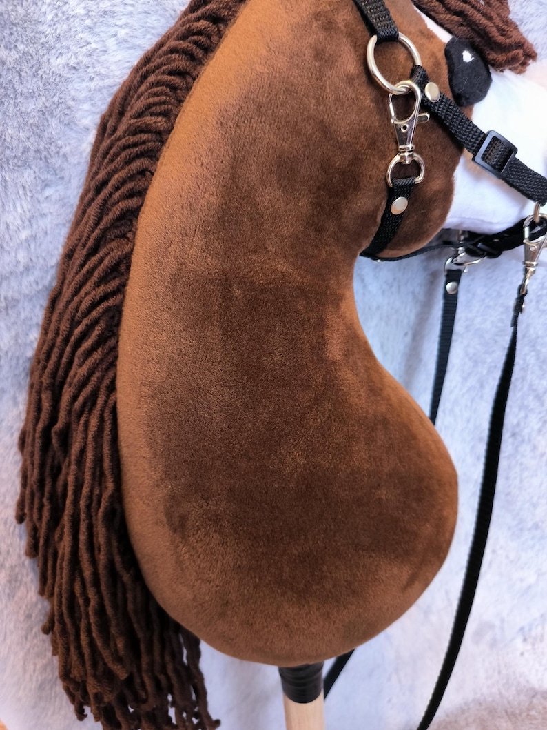 May include: A brown hobby horse with a plush head and a long, braided mane. The horse has a black bridle with silver-colored metal hardware and a wooden stick handle. The background is a light gray, textured fabric.