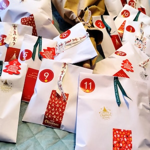 May include: A collection of white paper bags, each adorned with festive Christmas stickers and ribbons. Numbers 1 through 23 are visible on the bags, suggesting an advent calendar. Red and green accents add to the holiday theme.