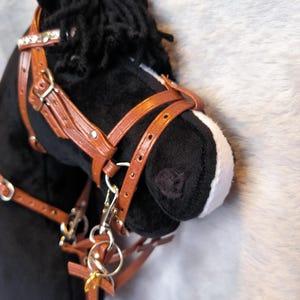 Könnte beinhalten: Ein schwarzer Plüsch-Steckenpferdkopf mit einem braunen Lederzaum und silberfarbenen Beschlägen. Das Pferd hat eine weiße Schnauze und eine schwarze Mähne. Ein dekoratives Strassband befindet sich am Zaum.