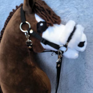 May include: A brown hobby horse head with a black bridle and reins. The horse has a white muzzle with black spots and a brown mane. The toy is made of soft fabric and is designed for children.
