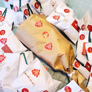 May include: An assortment of wrapped gifts, likely for an advent calendar. The gifts are wrapped in white and brown paper, adorned with red stickers featuring Christmas tree designs and numbered from 1 to 24. Green and red ribbons add festive accents.