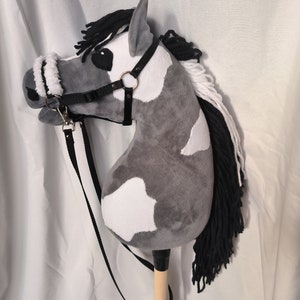 Könnte beinhalten: Ein grauer und weißer Plüschpferdekopf mit schwarzer Mähne und Schweif. Das Pferd trägt ein schwarzes Zaumzeug mit einem weißen Fleece-Nasenriemen und einem schwarzen Führseil.