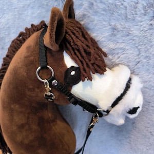 May include: A plush brown hobby horse head with a black bridle and reins. The horse has a white muzzle, black eyes, and a brown mane. The ears are brown and upright.