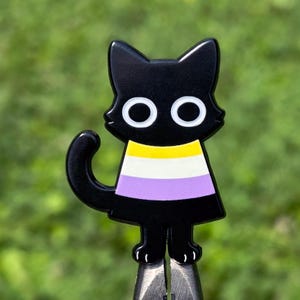 May include: A black cat enamel pin featuring large white eyes and a horizontal striped chest area with yellow, white, and purple bands. The cat has pointed ears, a curved tail, and small paws, with a smooth, glossy finish on the metal surface.