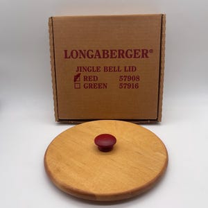May include: A wooden Longaberger Jingle Bell Lid with a red knob. The lid is round and light brown, with a darker brown rim. A brown cardboard box with the Longaberger logo and product information is behind the lid.