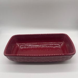 May include: A rectangular, burgundy-colored ceramic baking dish. The dish has a textured, basketweave-like pattern around the sides and a smooth interior. The dish is designed for baking and serving.