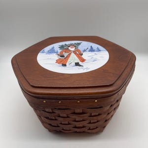 May include: A wooden basket with a woven base and a hexagonal lid. The lid features a circular illustration of Santa Claus carrying a Christmas tree in a snowy landscape. The basket is brown with brass-colored rivets.