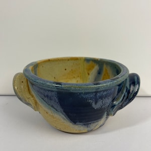 May include: A small, handmade ceramic bowl with two handles. The bowl is glazed in shades of blue and beige, with a speckled finish.
