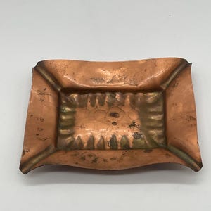 May include: A rectangular copper ashtray with a slightly curved shape. The ashtray has a hammered texture and a decorative design in the center. The edges are a darker shade of copper, and the overall color is a warm, reddish-brown.