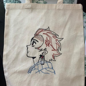 May include: A white canvas tote bag with an embroidered image of a character from the anime Demon Slayer. The character has red hair and is wearing a blue and white patterned shirt. The character is wearing a red and white earring with a sun design.