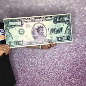 Puede incluir: Un gran billete de papel verde de un millón de dólares con una imagen de la Estatua de la Libertad. El billete tiene el texto "ONE MILLION DOLLARS" y "THE UNITED STATES OF AMERICA" impreso en él. El billete está siendo sostenido por la mano de una persona.