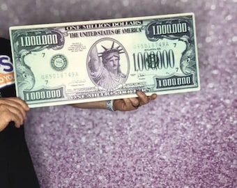 May include: A large, green, paper prop of a one million dollar bill featuring a picture of the Statue of Liberty. The bill has the text "ONE MILLION DOLLARS" and "THE UNITED STATES OF AMERICA" printed on it. The bill is being held by a person's hand.