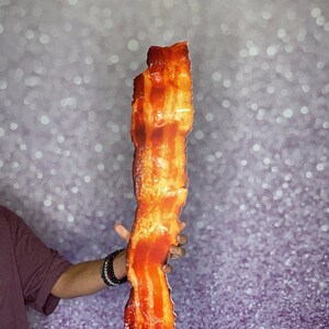 May include: A large, crispy, and realistic-looking bacon prop for photo booths. The prop is made of cardboard and is held by a person's hand. The text "Photo Booth Props By www.PropsToYou.com PROPS TO YOU" is visible at the bottom of the image.