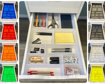 May include: A white drawer organizer with multiple compartments. The organizer is filled with various office supplies, including pens, pencils, scissors, a stapler, a ruler, and sticky notes. The drawer is open and the organizer is visible.