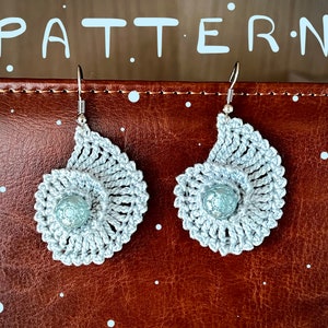 May include: A pair of silver crochet earrings with a spiral design and a light blue bead in the center. The earrings are hanging from silver hooks.