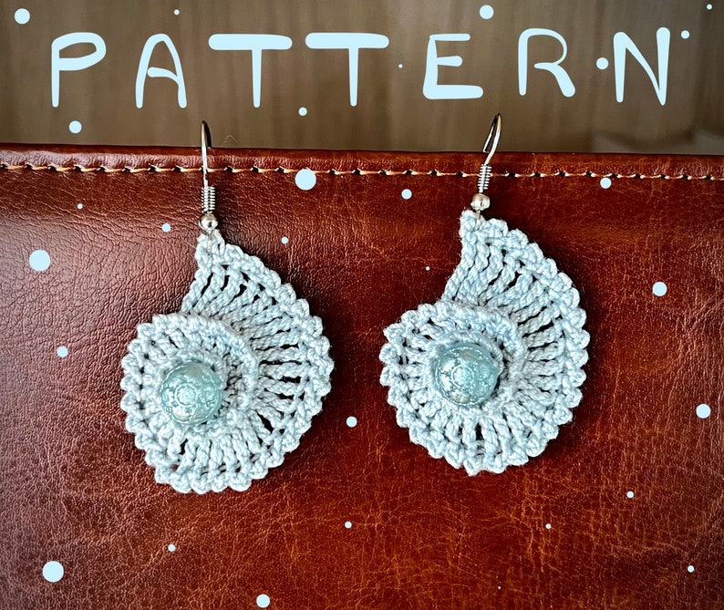 May include: A pair of silver crochet earrings with a spiral design and a light blue bead in the center. The earrings are hanging from silver hooks.