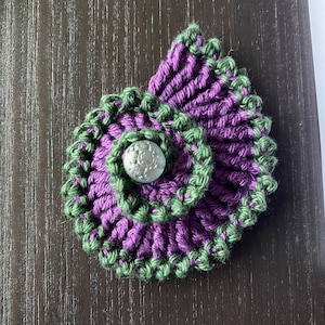 May include: A purple and green crocheted flower with a silver button in the center.