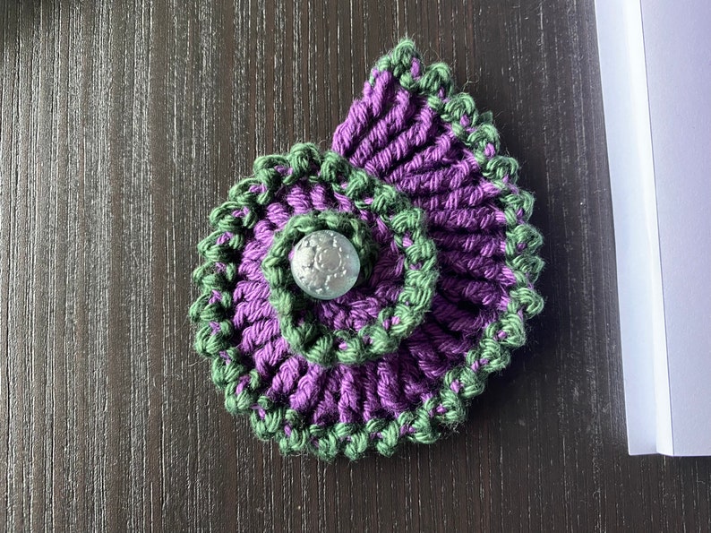 May include: A purple and green crocheted flower with a silver button in the center.