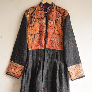 Peut inclure: Un manteau noir à manches longues doté d'un panneau supérieur orange vif brodé de motifs multicolores et d'incrustations de miroirs. La partie inférieure est noire avec des coutures géométriques rouges et blanches, un col haut et des boutons.