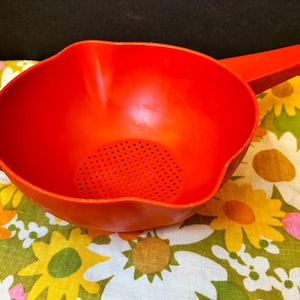 May include: A bright red plastic colander with a handle and a strainer at the bottom. The colander is sitting on a floral patterned tablecloth.
