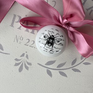 Puede incluir: Un adorno de cer&aacute;mica blanca con una ilustraci&oacute;n de abeja en blanco y negro y texto escrito a mano. El adorno est&aacute; atado con una cinta rosa.