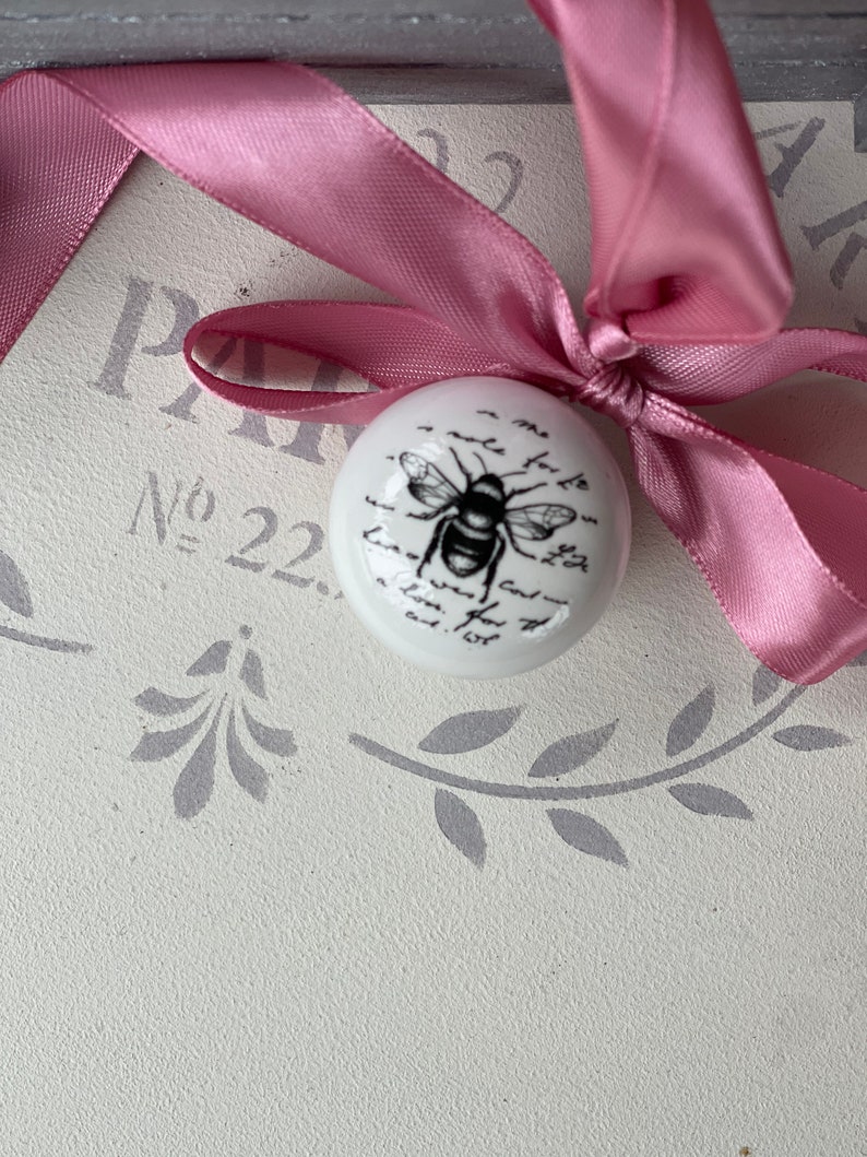 Puede incluir: Un adorno de cer&aacute;mica blanca con una ilustraci&oacute;n de abeja en blanco y negro y texto escrito a mano. El adorno est&aacute; atado con una cinta rosa.