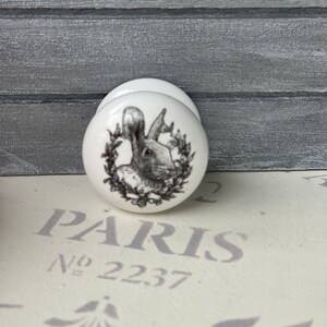 Puede incluir: Pomo de cer&aacute;mica blanco con una imagen en blanco y negro de un conejo en una corona. El pomo dice "PARIS No. 22.37."