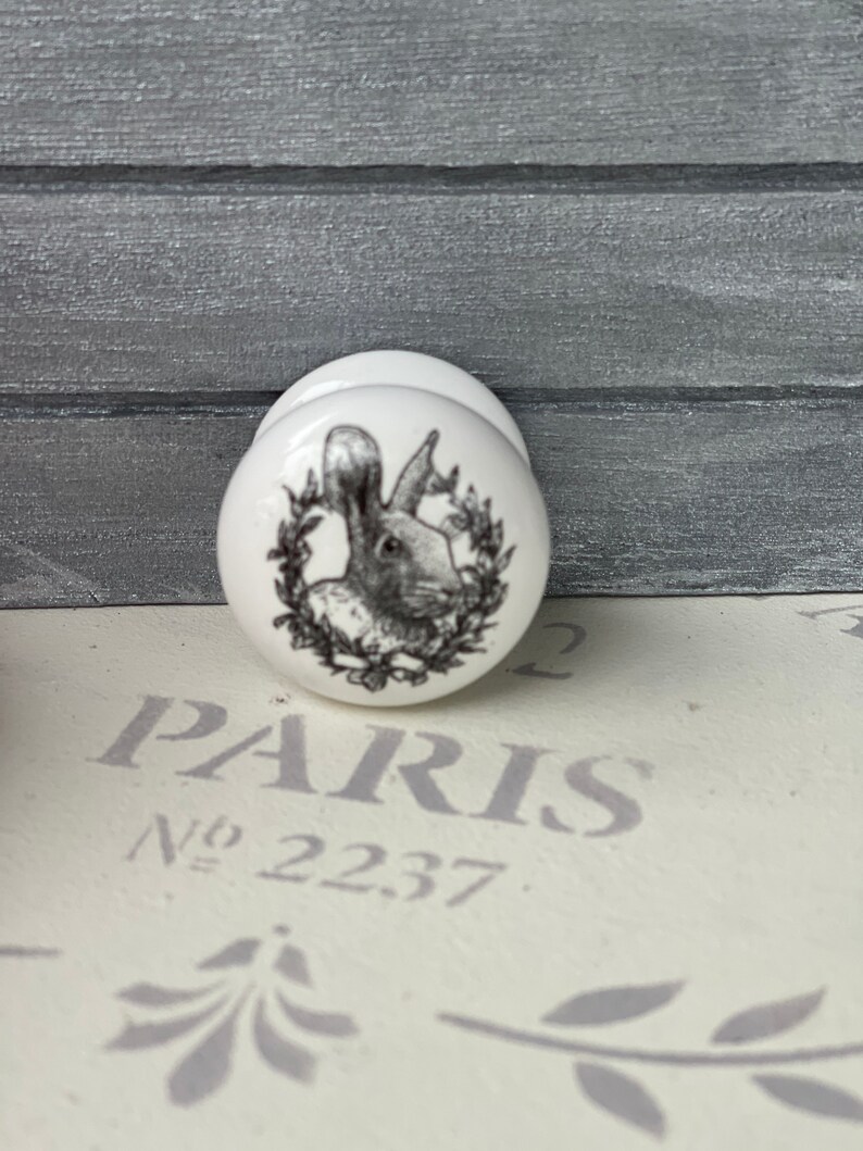 Puede incluir: Pomo de cer&aacute;mica blanco con una imagen en blanco y negro de un conejo en una corona. El pomo dice "PARIS No. 22.37."