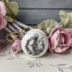 Puede incluir: Pomo de cer&aacute;mica blanco con una imagen en blanco y negro de un conejo en una corona. El pomo est&aacute; sobre una superficie blanca con un patr&oacute;n floral gris.